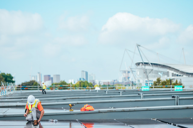 Workers installing solar panels on a rooftop with a city skyline in the background. The image includes construction workers wearing safety gear.