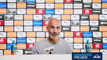 A man at a press conference table with a backdrop displaying various sponsor logos, flanked by two bottles.