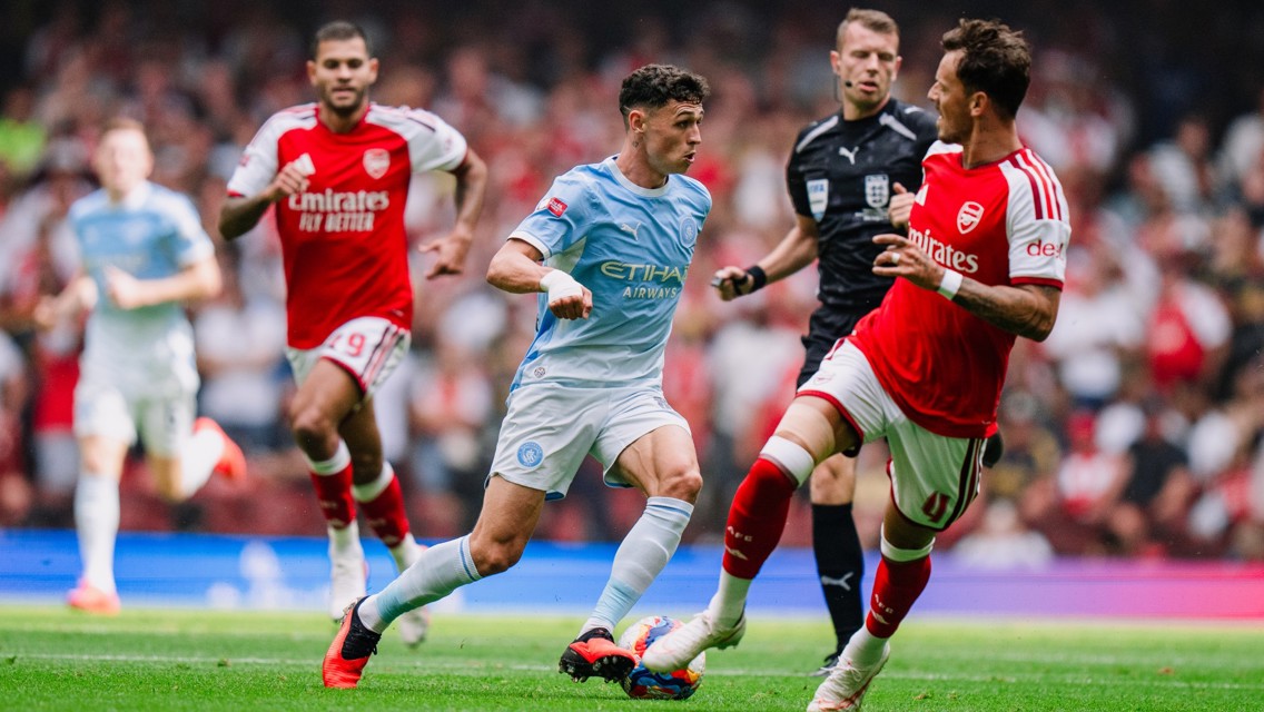A football match between players in blue Manchester City kit and red Arsenal kit with an official in black. One player in possession of the ball.