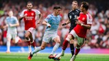 A football match between players in blue Manchester City kit and red Arsenal kit with an official in black. One player in possession of the ball.