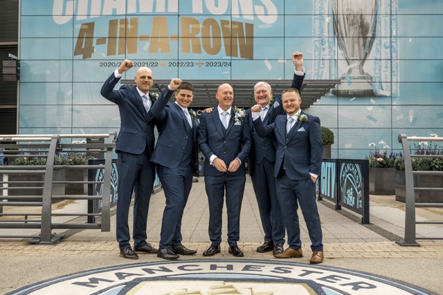 Six men in suits pose outside the Etihad Stadium in front of a Champions 4-in-a-row sign for the 2020/21, 2021/22, 2022/23, and 2023/24 seasons.