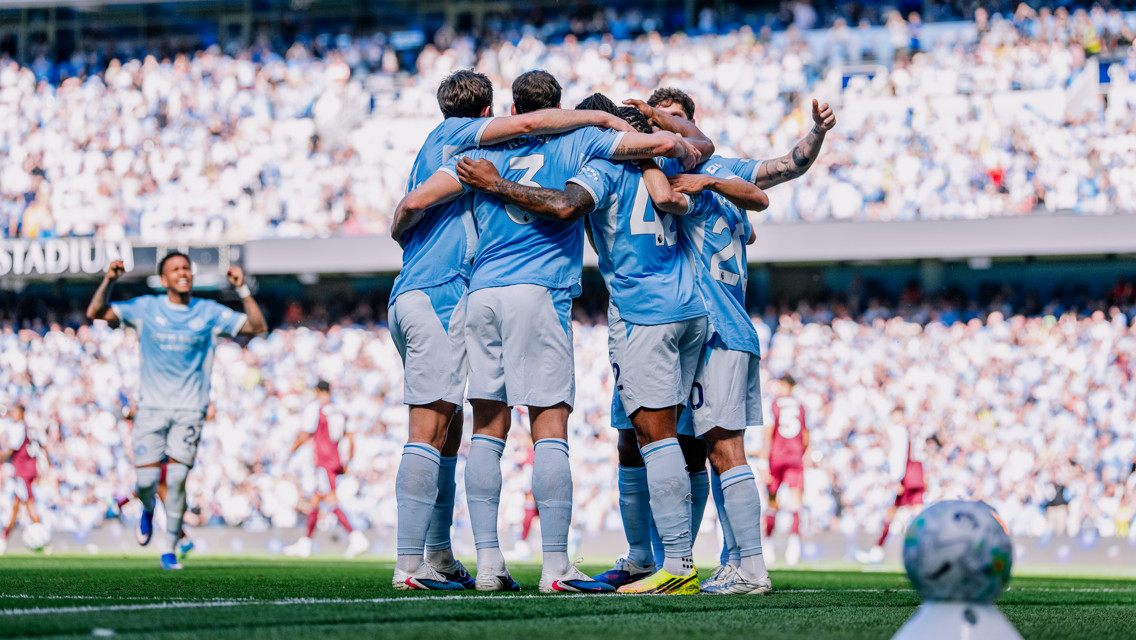 A group of football players in light blue jerseys, celebrating by huddling together on the field, surrounded by cheering fans at a stadium.