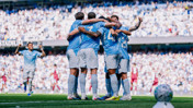 A group of football players in light blue jerseys, celebrating by huddling together on the field, surrounded by cheering fans at a stadium.