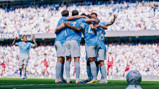 A group of football players in light blue jerseys, celebrating by huddling together on the field, surrounded by cheering fans at a stadium.