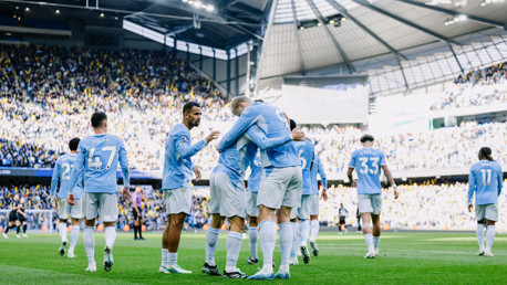 Manchester City players in light blue uniforms celebrating a goal at the Etihad Stadium.