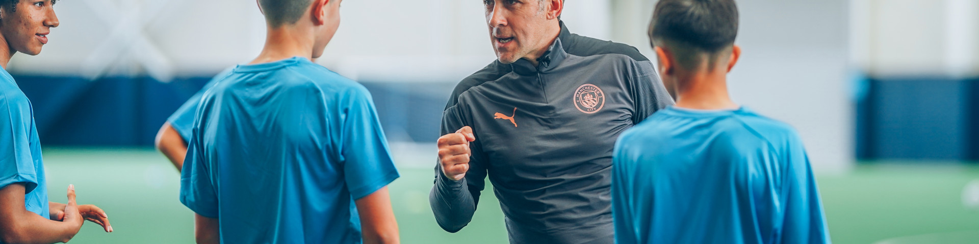 A coach instructs three young players wearing blue sportswear during a training session inside an indoor soccer facility. The coach wears a black jacket with a logo and gestures with a clenched fist.