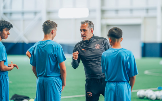 A coach instructs three young players wearing blue sportswear during a training session inside an indoor soccer facility. The coach wears a black jacket with a logo and gestures with a clenched fist.