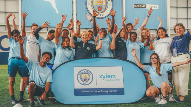 A group of people in light blue t-shirts with 'Cityzens Giving' logo, standing in front of a Manchester City and Xylem promotional banner inside a football facility.