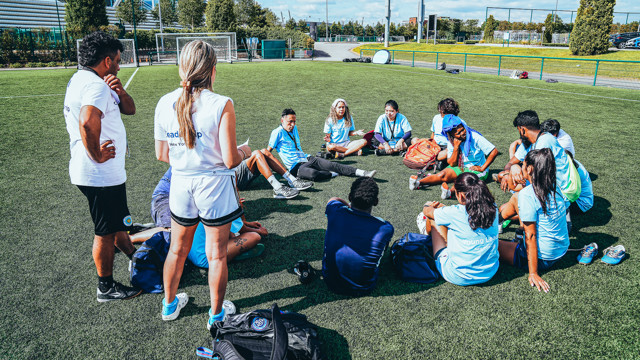 A group of young soccer players sits in a circle with their coaches on a field listening to a discussion or training instructions.