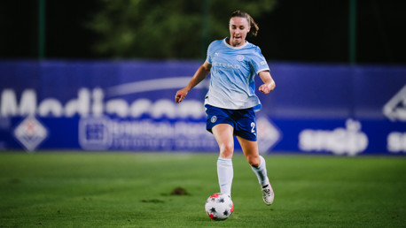A female soccer player in a Manchester City uniform running with the ball on a field.