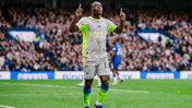 A Manchester City player in a grey and neon yellow kit celebrates on the field with raised arms. The player has the number 11 on their shorts. The background shows a stadium crowd.