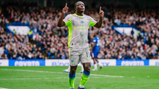 A Manchester City player in a grey and neon yellow kit celebrates on the field with raised arms. The player has the number 11 on their shorts. The background shows a stadium crowd.