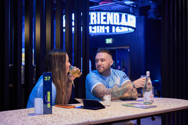 Two people wearing Manchester City jerseys talking at a bar table. The setting has a blue neon light, table number, and drinks.