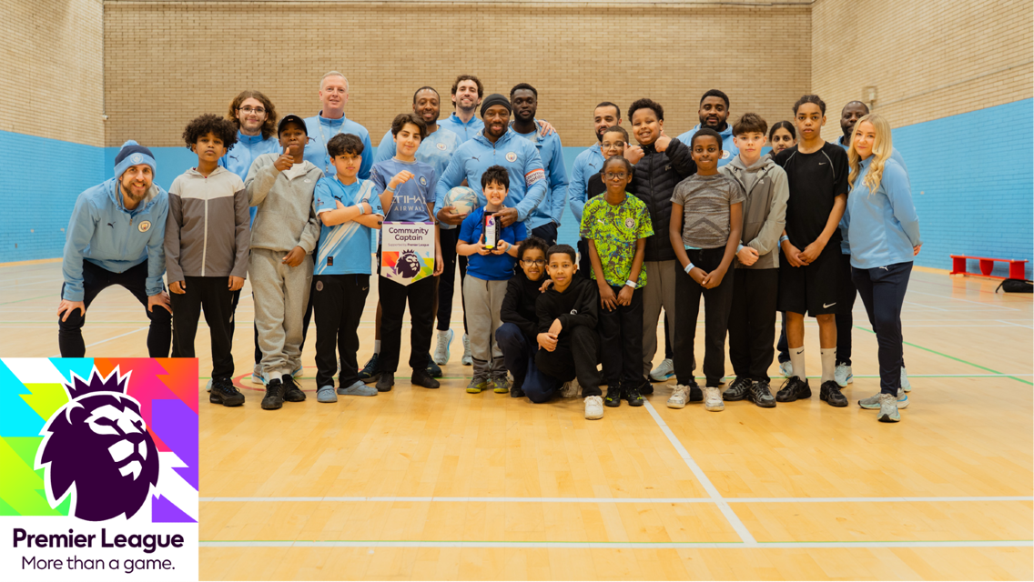 Group photo of adults and children in a sports hall, with several wearing Manchester City attire, and a Premier League banner displayed in the foreground.