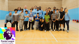 Group photo of adults and children in a sports hall, with several wearing Manchester City attire, and a Premier League banner displayed in the foreground.