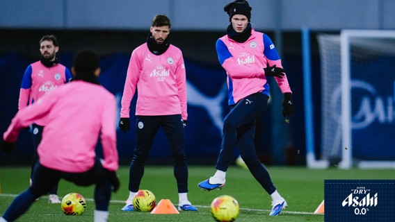 Manchester City players in pink training gear during a practice session on a field with cones and footballs, featuring Asahi Super Dry 0.0% logo.