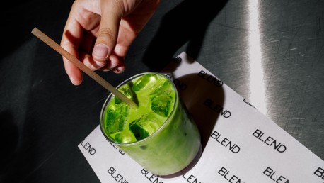 Hand stirring a green matcha iced drink in a clear glass on a surface labeled 'BLEND'.