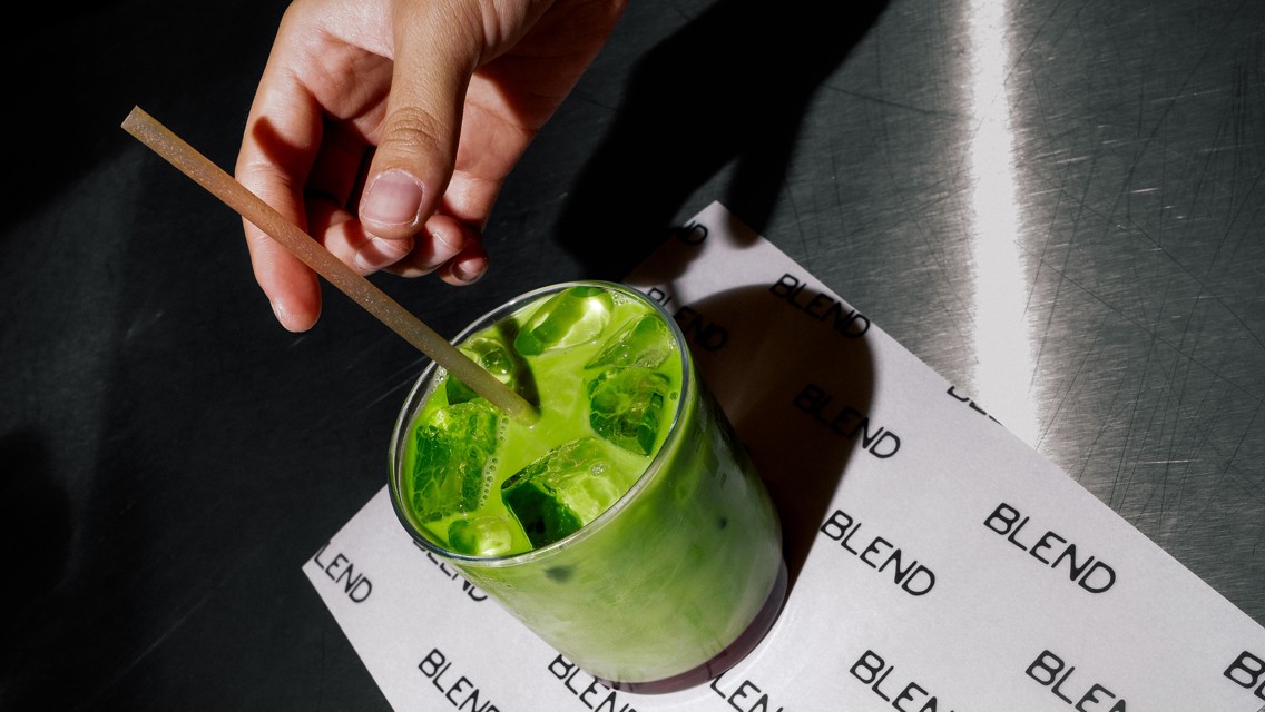Hand stirring a green matcha iced drink in a clear glass on a surface labeled 'BLEND'.