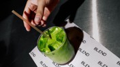 Hand stirring a green matcha iced drink in a clear glass on a surface labeled 'BLEND'.