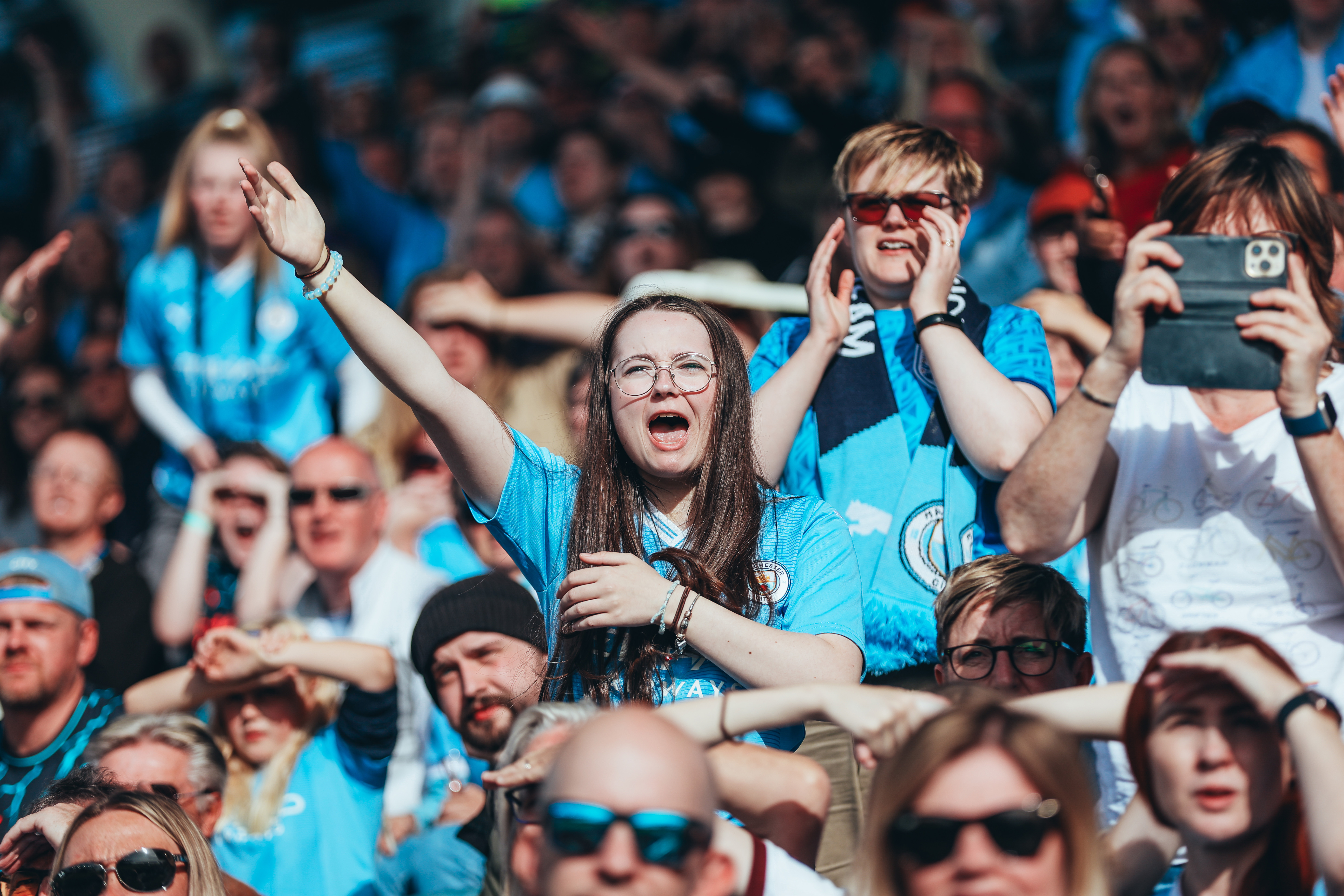 Fans in light blue Manchester City jerseys cheer in a stadium. Faces are blurred, and one fan holds an arm up while another takes a photo.