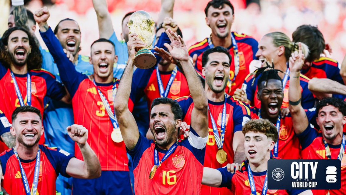 A group of football players in red and navy jerseys celebrating with a trophy. The team is jubilant, with players raising their hands and smiling widely. The image includes a logo related to a World Cup and Manchester City.