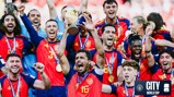 A group of football players in red and navy jerseys celebrating with a trophy. The team is jubilant, with players raising their hands and smiling widely. The image includes a logo related to a World Cup and Manchester City.