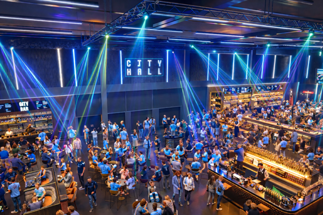 A lively indoor venue with people wearing blue jerseys, enjoying drinks and socializing under colorful lights. Signage includes 'City Hall' and 'City Bar'.
