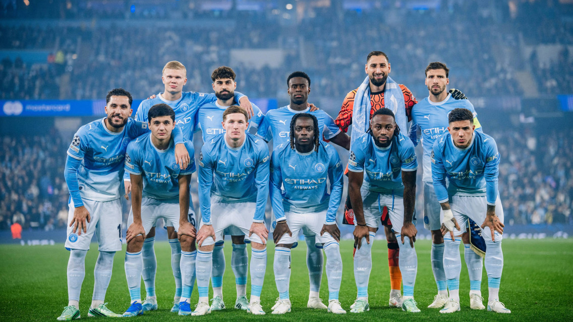 Manchester City football team posing for a group photo in blue jerseys at a stadium.