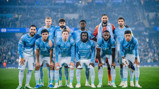 Manchester City football team posing for a group photo in blue jerseys at a stadium.