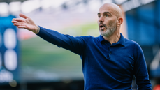 A bald man in a blue shirt makes a gesturing motion, indicative of coaching or giving directions, on a blurred sports field.