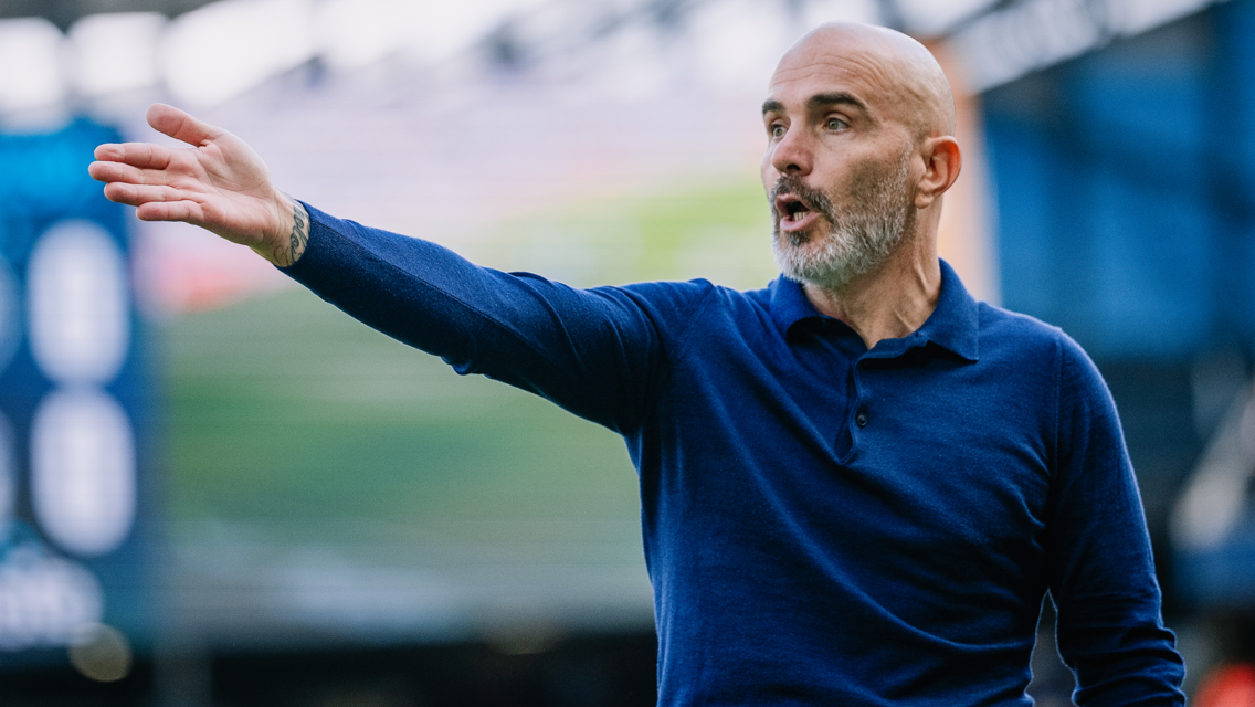 A bald man in a blue shirt makes a gesturing motion, indicative of coaching or giving directions, on a blurred sports field.