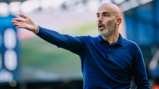 A bald man in a blue shirt makes a gesturing motion, indicative of coaching or giving directions, on a blurred sports field.