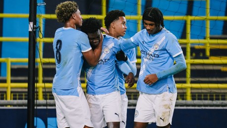 Manchester City players wear light blue uniforms celebrating together, standing on a football field. The team's official badge is visible on their shirts.