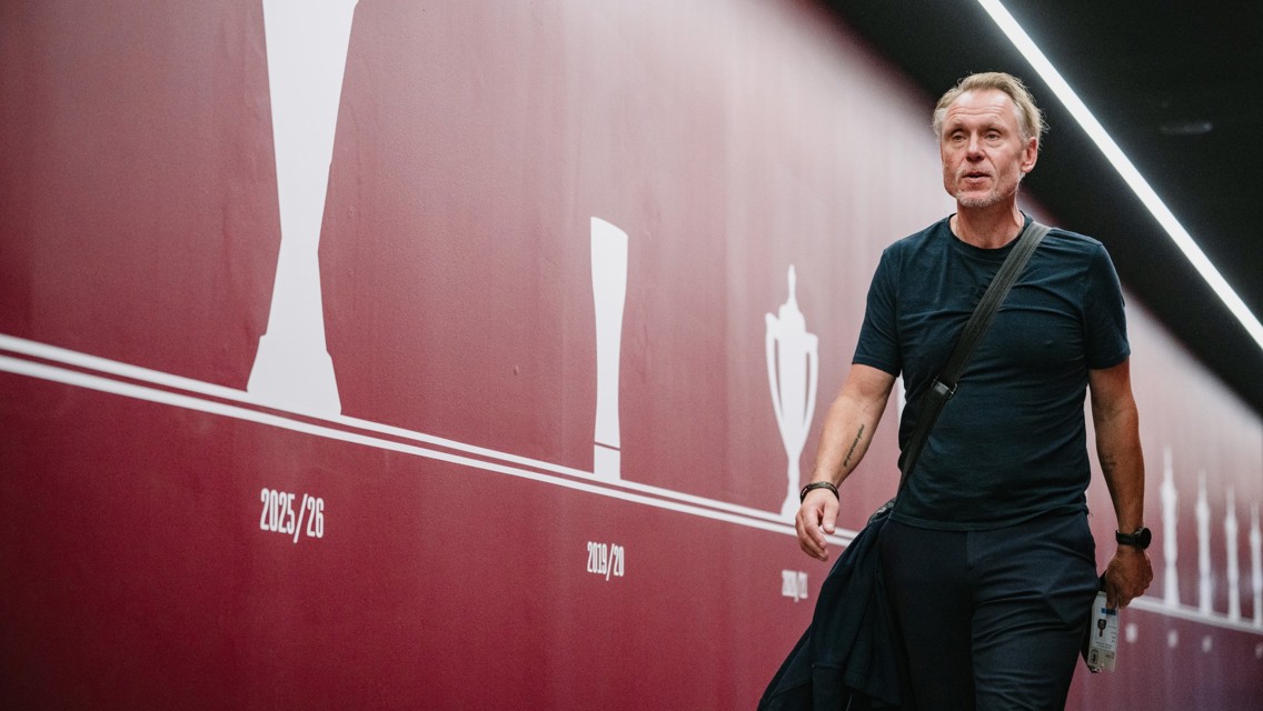 A man in a dark outfit walks through a red sports tunnel decorated with trophy silhouettes and dates like '2025/26' and '2019/20.'