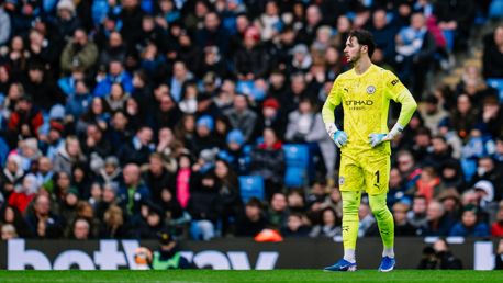 A goalkeeper wearing a Man City kit stands on the field with spectators in the background.