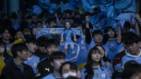 Blurred image of excited fans wearing Manchester City jerseys waving flags and holding a plush toy in a crowd setting.