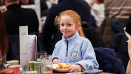 Young girl wearing a Manchester City jacket sitting at a table with a slice of pizza at an event.