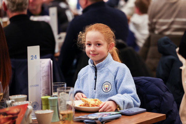 Young girl wearing a Manchester City jacket sitting at a table with a slice of pizza at an event.