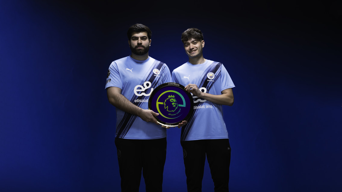 Two people in Manchester City esports jerseys holding an ePremier League champion trophy.