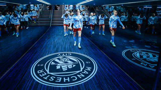 Manchester City women players walk down the tunnel, with their logo visible on the floor, reflecting in mirrored walls.