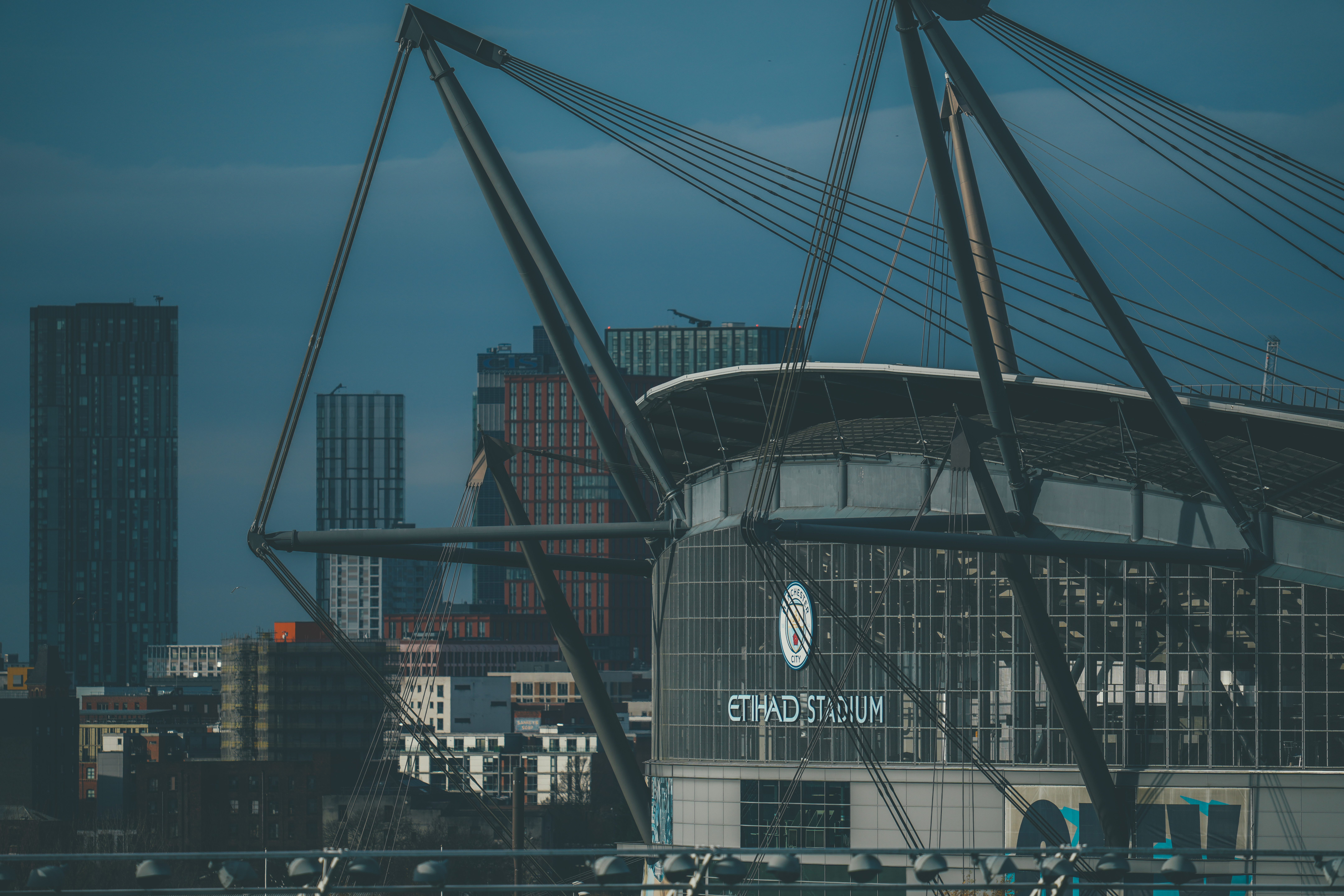 A view of the Etihad Stadium with buildings in the background. The stadium is partially obscured by structural elements.