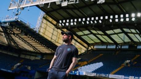 A person in a cap and sportswear is standing inside a stadium, with empty seats and a large roof structure visible in the background.