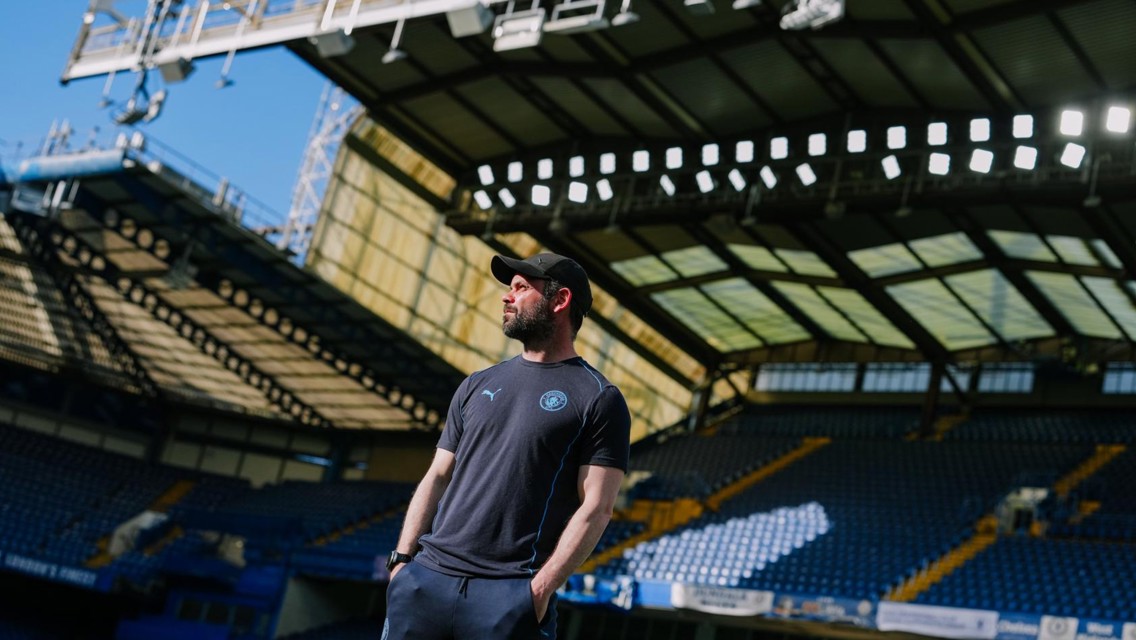 A person in a cap and sportswear is standing inside a stadium, with empty seats and a large roof structure visible in the background.