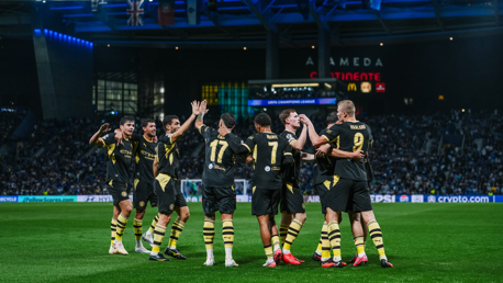 A group of Manchester City players celebrating on the field during a UEFA Champions League match. They are wearing black and yellow kits with numbers visible on their backs.