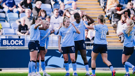 Players from Manchester City Women's Team celebrate on the field wearing their light blue jerseys, watched by a crowd in the stadium.