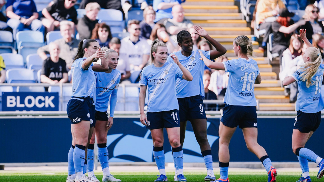 Players from Manchester City Women's Team celebrate on the field wearing their light blue jerseys, watched by a crowd in the stadium.