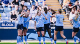 Players from Manchester City Women's Team celebrate on the field wearing their light blue jerseys, watched by a crowd in the stadium.