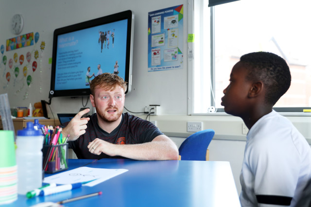A teacher is explaining something to a student in a classroom setting. The background includes a digital screen displaying a presentation and various classroom decorations.