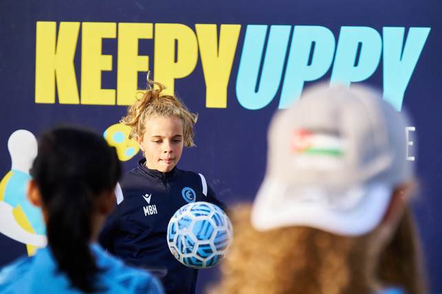 A young child with curly blonde hair playing with a football, wearing sports gear with 'MBH' initials. The background visible includes a 'KEEPY UPPY' banner, with other individuals partially visible.