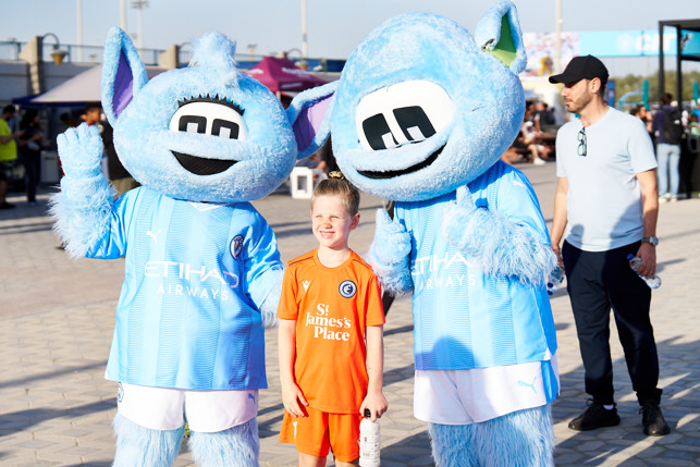 Two Manchester City mascots, Moonchester and Moonbeam, pose with a young fan holding a water bottle at a fan event.